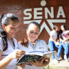 Dos estudiantes del SENA mirando un cuaderno con una pared con el logo del SENA en el fondo.