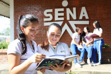 Dos estudiantes del SENA mirando un cuaderno con una pared con el logo del SENA en el fondo.