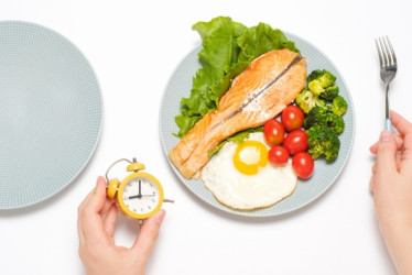 Persona sentada frente a un plato de comida sosteniendo un reloj en su mano izquierda.